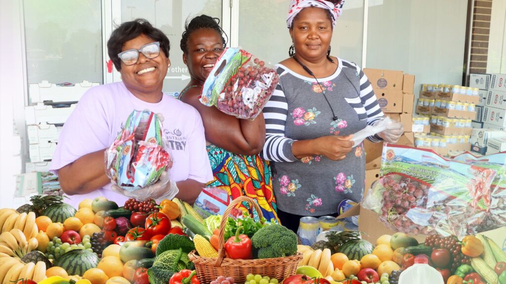 Food pantry volunteers smile behind a table full of fresh fruit and vegetables