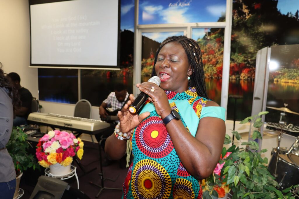 A worship leader in a colorful African print dress sings into a microphone at Faith Klinik